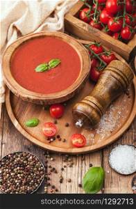 Wooden plate of creamy tomato soup 0n round tray, pepper and kitchen cloth on wooden board with box of raw tomatoes.