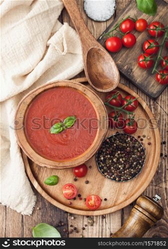 Wooden plate of creamy tomato soup 0n round tray, pepper and kitchen cloth on wooden board with box of raw tomatoes. Top view