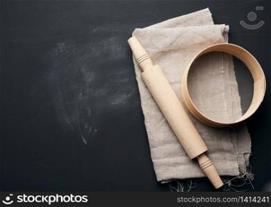 wooden rolling pin and a round sieve lie on a gray linen napkin, black table, top view, copy space
