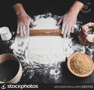 wooden rolling pin in male hands and ingredients on a black wooden table