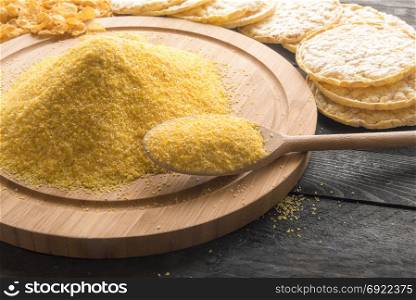 Wooden spoon full of corn flour on a round wooden board, surrounded by products made from it, corn flakes and puffed corn cakes, on a vintage table, in the sunlight.