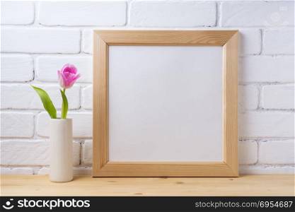 Wooden square picture frame mockup with pink tulip in cylinder vase near painted brick wall. Empty frame mock up for presentation design. Template framing for modern art.