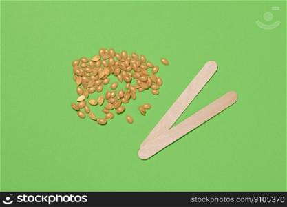 Wooden sticks for applying wax and yellow wax granules for cosmetic procedures on a green background, top view