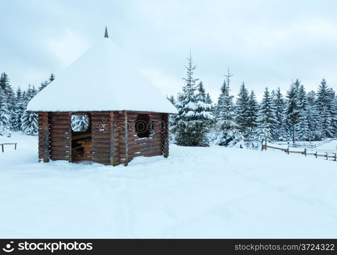 Wooden summerhouse on winter hill top and fir forest behind . Cloudy day.