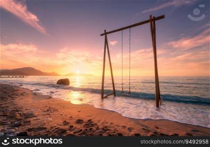 Wooden swing in beautiful blue sea with waves, sandy beach, purple sky with golden sunlight at sunset in summer. Vacation in Oludeniz, Turkey. Tropical landscape with swing on sea coast, water. Travel