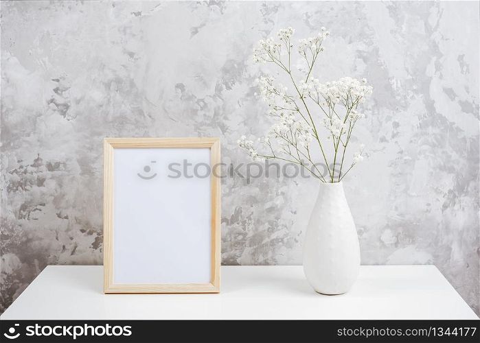 Wooden vertical white empty frame and bouquet of white small flowers gypsophila in vase on table on gray concrete wall background. Mockup Template for your design, text.. Wooden vertical white empty frame and bouquet of white small flowers gypsophila in vase on table on gray concrete wall background. Mockup Template for your design, text