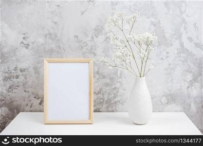 Wooden vertical white empty frame and bouquet of white small flowers gypsophila in vase on table on gray concrete wall background. Mockup Template for your design, text.. Wooden vertical white empty frame and bouquet of white small flowers gypsophila in vase on table on gray concrete wall background. Mockup Template for your design, text