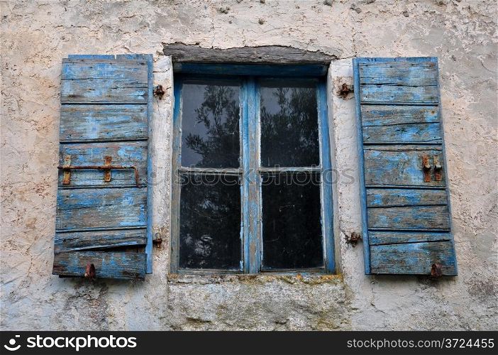 Wooden window shutter with chipped blue paint and textured wall of old house.