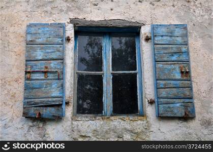 Wooden window shutter with chipped blue paint and textured wall of old house.