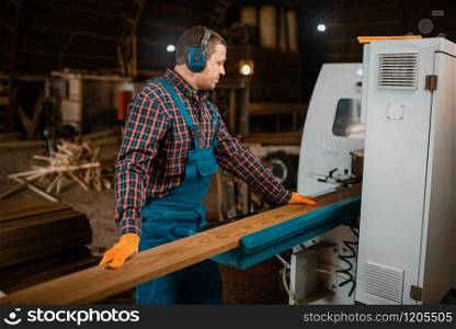 Woodworker in uniform and headphones works on woodworking machine, lumber industry, carpentry. Wood processing on factory