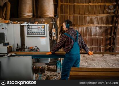 Woodworker in uniform and headphones works on woodworking machine, lumber industry, carpentry. Wood factory. Woodworker works on machine, lumber industry
