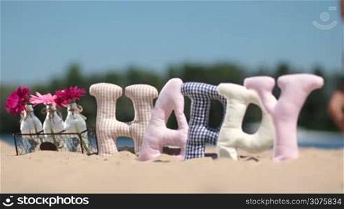 Word happy made of fabric padded letters and flowers in glass bottles on the sand at the beach over blurred background of kid running on shore.