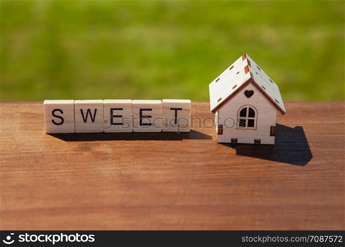 Word sweet of wooden letters and small toy wooden house on brown surface, green grass in background. Concept sweet home, mortgage, dream house, real estate acquisition. Soft selective focus.