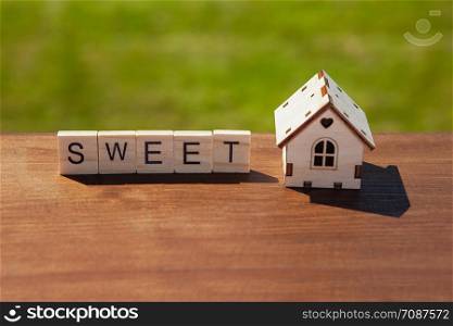 Word sweet of wooden letters and small toy wooden house on brown surface, green grass in background. Concept sweet home, mortgage, dream house, real estate acquisition. Soft selective focus.