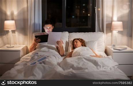 work, deadline and people concept - man with tablet computer and papers working in bed at night. man with tablet computer working in bed at night