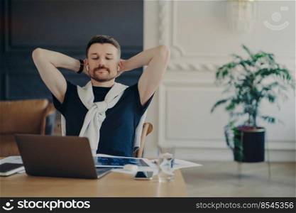 Work done. Relaxed man freelancer or enterpruer in wireless earphones sitting at table with hands behind head, watching business webinar or listening speaker on laptop while working remotely from home. Young businessman watching video on laptop and feeling relaxed while working from home