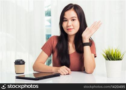 Work from home, Asian business young beautiful woman smiling sitting on desk workspace raise hand say hello communicating with team video chat or conferencing on digital tablet computer at home office