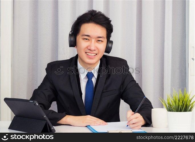 Work from home, Asian young businessman wear suit video conference call or facetime he smiling sitting on desk using smart digital tablet computer and write paper notebook information at home office