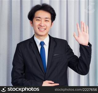 Work from home, Close up face of Asian young businessman wear suit video conference call or facetime he looking to camera and raise your hand and say hello team or customer at the home office