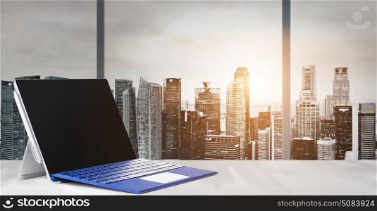 Work place with panoramic view. Laptop on table in office with panoramic sunset view of modern downtown skyscrapers at business district, blank screen