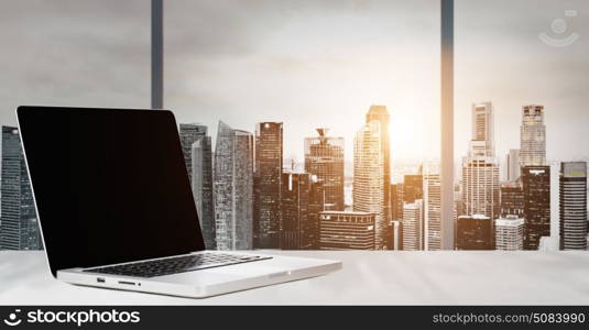 Work place with panoramic view. Laptop on table in office with panoramic sunset view of modern downtown skyscrapers at business district, blank screen