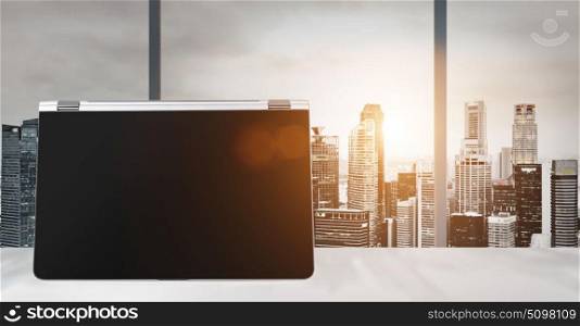 Work place with panoramic view. Laptop on table in office with panoramic sunset view of modern downtown skyscrapers at business district, blank screen