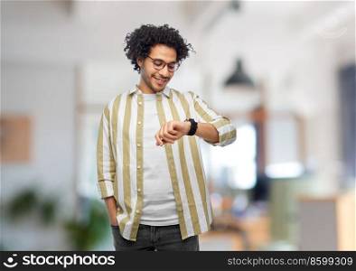 work, technology and people concept - smiling young man in glasses with smart watch over office background. smiling man in glasses with smart watch at office
