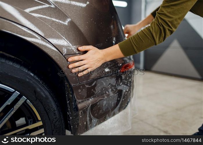 Worker applies car protection film on rear bumper. Installation of coating that protects the paint of automobile from scratches. New vehicle in garage, tuning procedure. Worker applies car protection film on rear bumper
