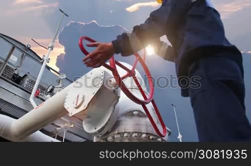 worker closes ball valve on cooling installations at gas compressor station, against background of sunset, closeup