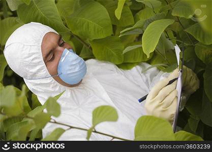 Worker in protective mask and suit writing on pad amongst plants