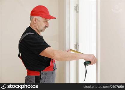 Worker in red cap and work suit with the measuring tape. Man is installing the doors. Measure tape in hands. Repair works. Maintenance in the apartment. Worker in red cap and work suit with the measuring tape. Man is installing the doors. Measure tape in hands. Repair works. Maintenance in the apartment.