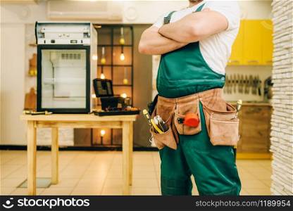 Worker in uniform against refrigerator on the table at home. Repairing of fridge occupation, professional service