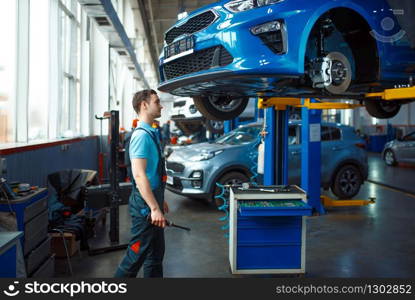 Worker in uniform checks vehicle bottom on lift, car service station. Automobile checking and inspection, professional diagnostics and repair. Worker in uniform checks vehicle bottom on lift