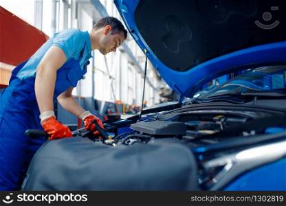 Worker in uniform checks vehicle engine, car service station. Automobile checking and inspection, professional diagnostics and repair. Worker in uniform checks engine, car service