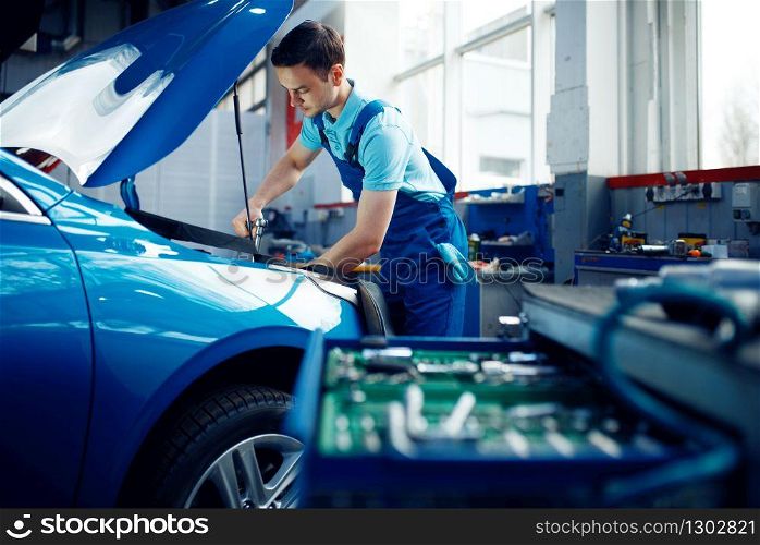 Worker in uniform checks vehicle engine, car service station. Automobile checking and inspection, professional diagnostics and repair. Worker in uniform checks engine, car service