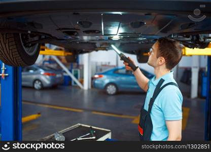Worker in uniform checks vehicle suspension on lift, car service station. Automobile checking and inspection, professional diagnostics and repair. Worker checks vehicle suspension, car service