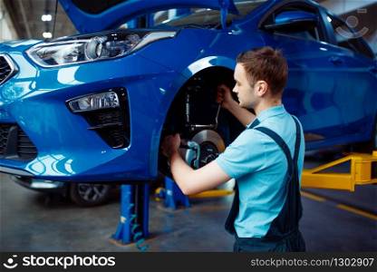Worker in uniform fix problem on vehicle with removed wheel, car service station. Automobile checking and inspection, professional diagnostics and repair. Worker fix problem on vehicle, car service