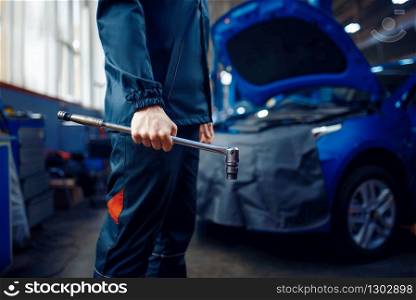 Worker in uniform holds wrench, vehicle with opened hood on background, car service station. Automobile checking and inspection, professional diagnostics and repair. Worker in uniform holds wrench, car service