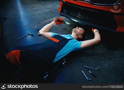 Worker in uniform lying under the vehicle, car service station. Automobile checking and inspection, professional diagnostics and repair. Worker lying under the vehicle, car service