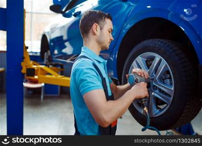 Worker in uniform removes wheel from vehicle on lift, car tire service station. Automobile checking and inspection, professional diagnostics and repair. Worker removes wheel from vehicle on lift