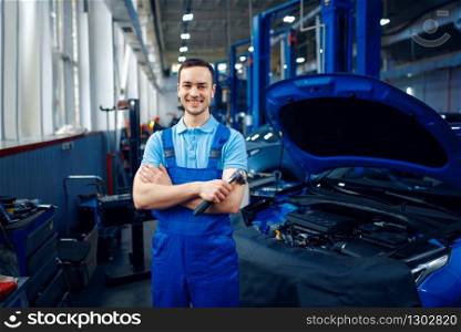 Worker in uniform stands at vehicle on lift, car service station. Automobile checking and inspection, professional diagnostics and repair. Worker stands at vehicle on lift, car service