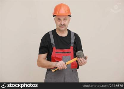 worker man in uniform, overalls and protective helmet is holding rubber hammer with yellow handle on white background.. worker man in uniform, overalls and protective helmet is holding rubber hammer with yellow handle on white background