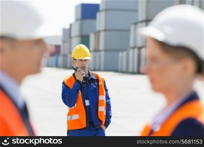 Worker using walkie-talkie while colleagues discussing in foreground at shipping yard