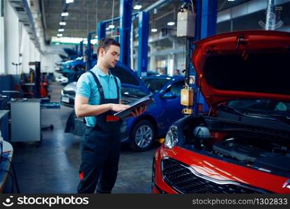 Worker with a checklist stands at vehicle with opened hood, car service station. Automobile checking and inspection, professional diagnostics and repair. Worker with a checklist, car service station