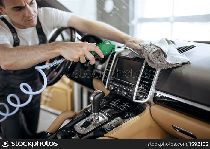 Worker with air gun cleans car air duct grating, dry cleaning and detailing. Vehicle washing in garage, thoroughly care of automobile. Worker with air gun cleans car air duct grating