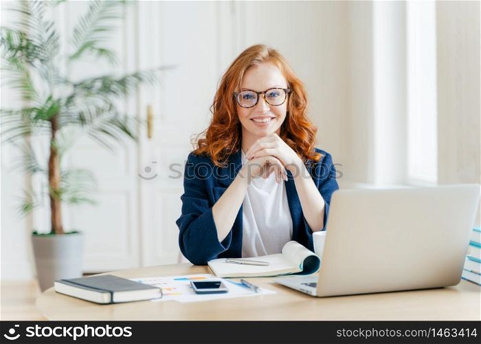 Working concept. Intelligent manager with satisfied expression, writes in notepad, works on modern laptop computer, uses organizer app, sits against office interior. Experienced entrepreneur