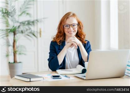 Working concept. Intelligent manager with satisfied expression, writes in notepad, works on modern laptop computer, uses organizer app, sits against office interior. Experienced entrepreneur