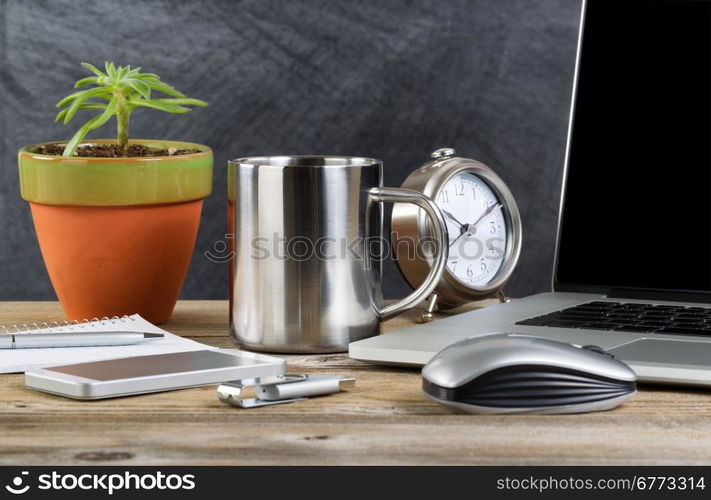 Working desktop with laptop, mouse, clock, cell phone, notebook, pen, thumb drive, plant and coffee mug in front of blackboard. Layout in horizontal format with copy space.