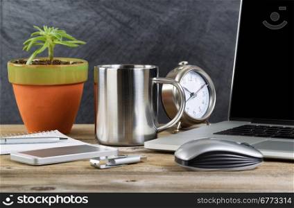 Working desktop with laptop, mouse, clock, cell phone, notebook, pen, thumb drive, plant and coffee mug in front of blackboard. Layout in horizontal format with copy space.