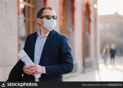 Working in pandemic situation. Business worker or entrepreneur wears medical mask for coronavirus protection stands beside office building outdoor holds newspapers concentrated somewhere into distance